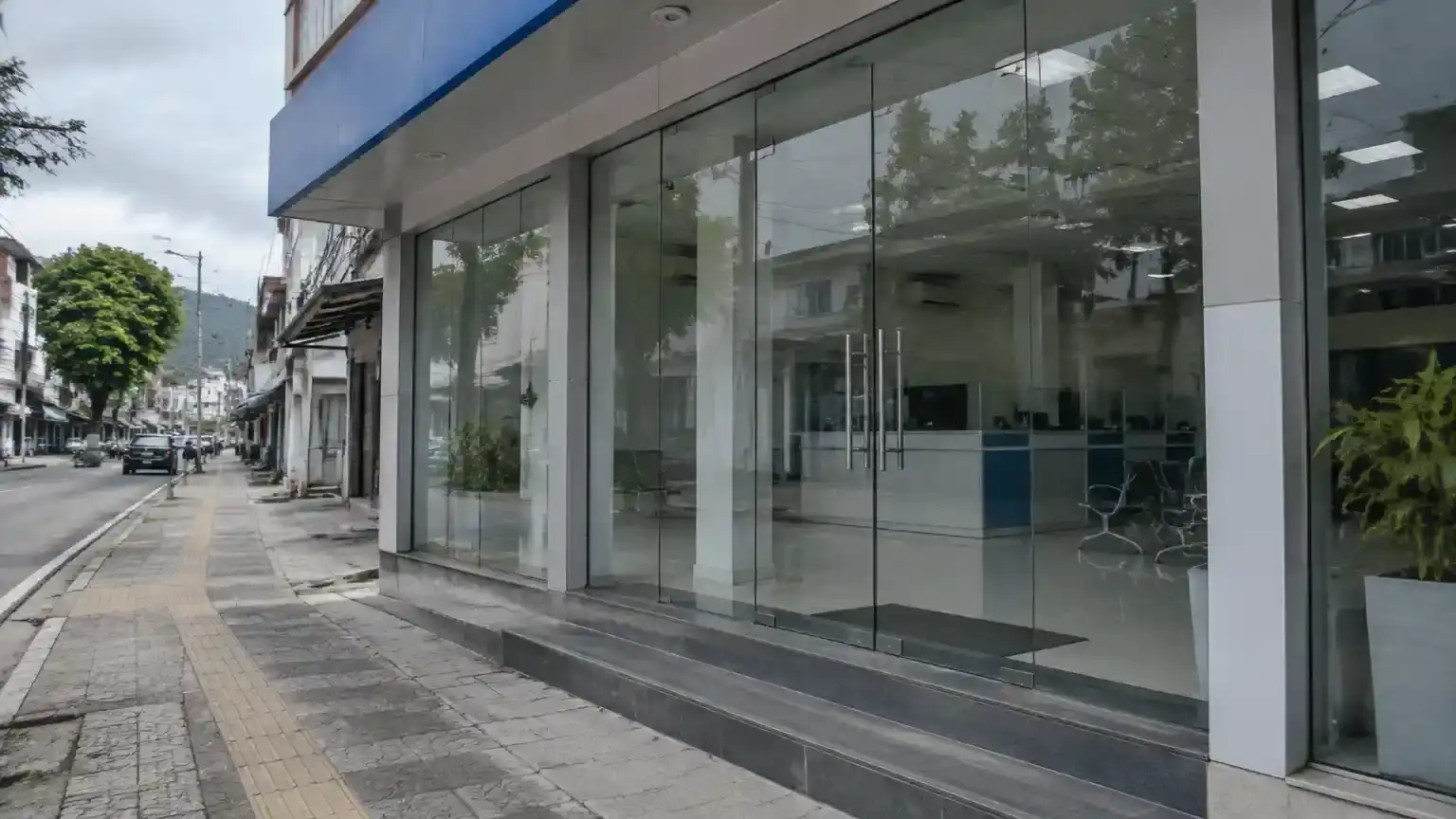 Kantor bank kecil generik di Indonesia tampak tidak beroperasi. Pintu kaca tertutup, sementara ruang layanan yang kosong terlihat dari trotoar dalam suasana siang yang tenang. Tampak luar kantor bank kecil dengan pintu kaca tertutup dan ruang layanan kosong.